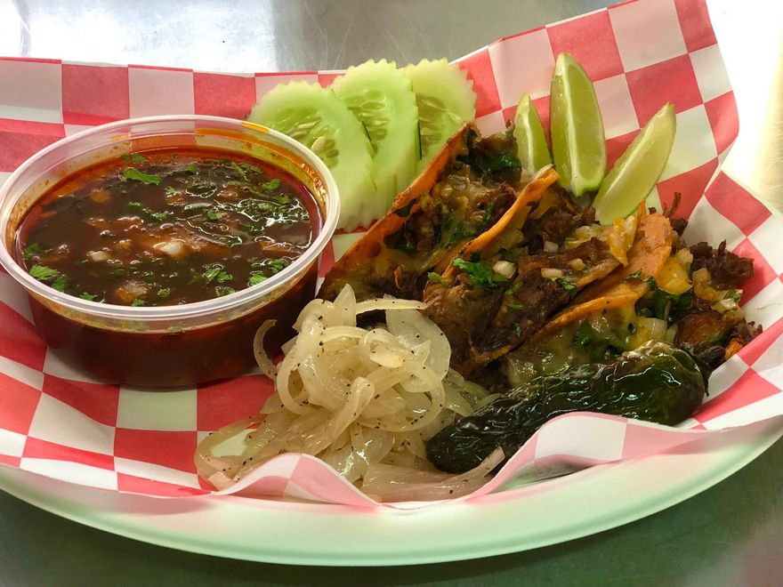 Birria tacos served with a cup of consommé, grilled onions, jalapeño, cucumber, and lime.